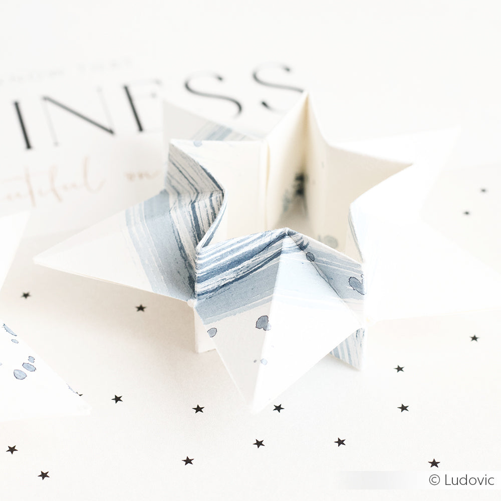 Ein weiß und blau marmorierter Papierstern von Alexandra Renke, hergestellt mit Designpapieren und dem Motivstanze Doppel-Faltstern DieCut System, ruht auf einer weißen Fläche, die mit schwarzem Sternenkonfetti bestreut ist.