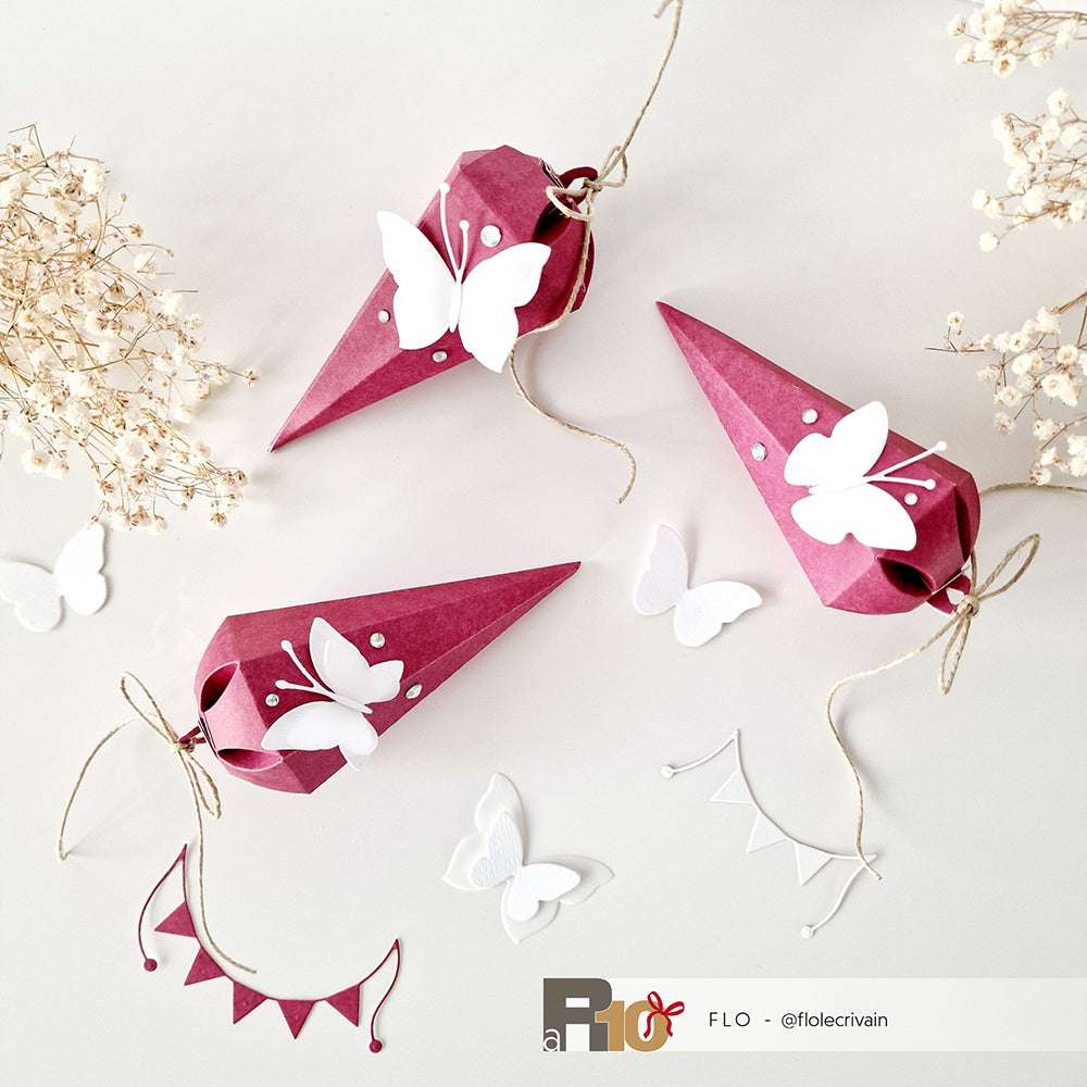 Drei rosafarbene, mit weißen Schmetterlingen verzierte Papiertüten, die mit der Motivstanze Schultüte und Designpapieren von Alexandra Renke gebastelt wurden, sind zwischen Trockenblumen und Papierausschnitten angeordnet.