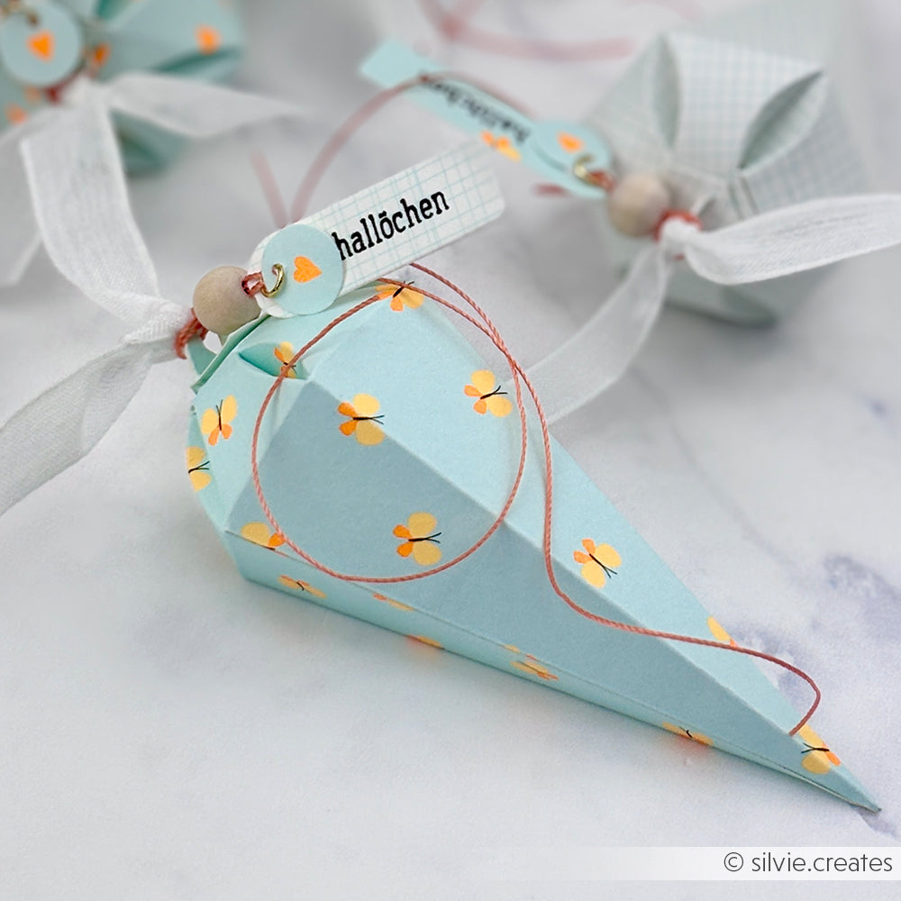 Hellblauer Papierkegel mit Blumenmuster, gefertigt mit Designpapier-Stanzformen und verziert mit einem Band, einer Perle und einem kleinen Anhänger mit der Aufschrift „hallöchen“, präsentiert auf einem hellen Untergrund.