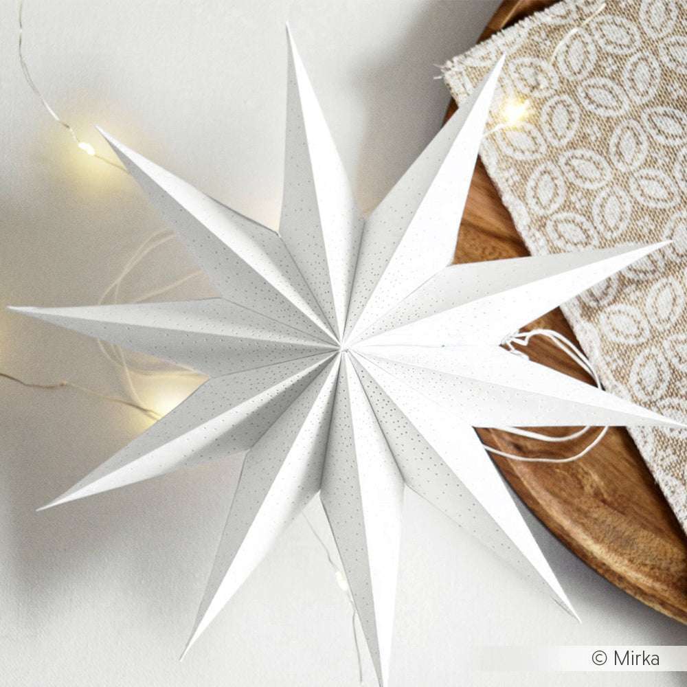 Ein weißer Papierstern von Alexandra Renke, gebastelt mit Designpapieren und einer Motivstanze Faltstern, steht auf einem Tisch, der mit Lichterketten und einem gemusterten beigen Tuch geschmückt ist.