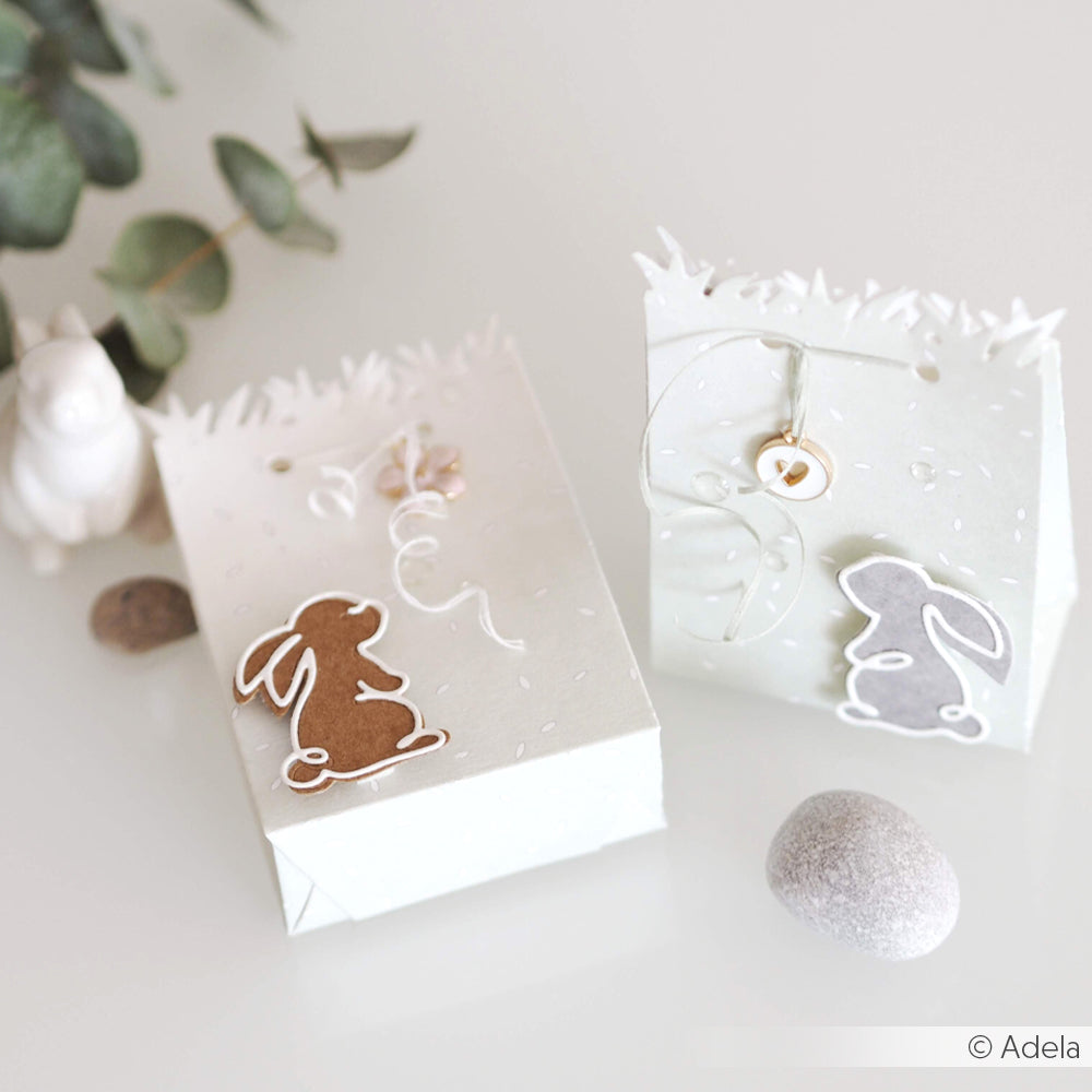 Zwei weiße Geschenktüten, die mit Alexandra Renke Designpapieren und der Motivstanze Tüte Kleine Wiese gestaltet wurden, sind mit Hasenausschnitten, Schleifenband und Kieselsteinen verziert und bieten ein zartes Frühlingsmotiv.