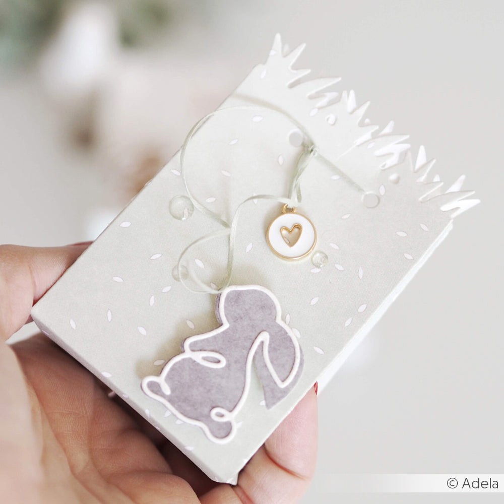 Eine Hand hält eine niedliche Hasen-Geschenkbox aus Alexandra Renke Designpapieren, die mit einem Herzanhänger und weißen Akzenten verziert ist und mit der Alexandra Renke Motivstanze Tüte Kleine Wiese veredelt wurde.