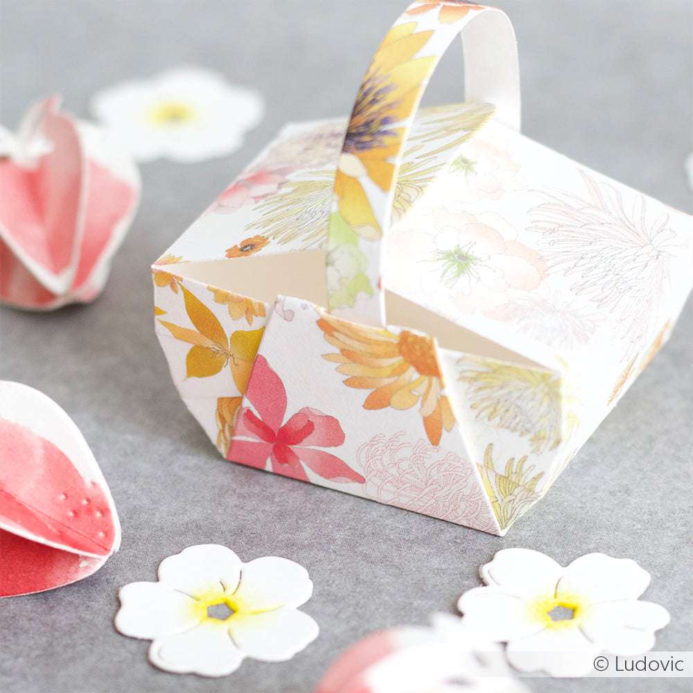 Ein kleiner, floral gemusterter Papierkorb, gefertigt mit Alexandra Renke Designpapieren und einer Motivstanze Picknickkorb, umgeben von Papierblumen auf einer grauen Oberfläche.