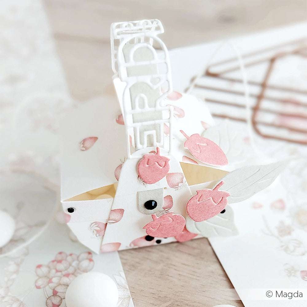 Eine dekorative Geschenkbox aus Blumenpapier von Alexandra Renke mit rosa Papier-Erdbeeren und einem weißen HAPPY-Topper - perfekt für jeden Anlass.