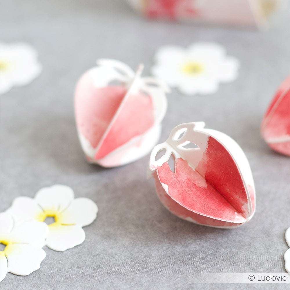 Mit der Alexandra Renke Motivstanze Erdbeere aus hochwertigen Designpapieren gestaltete Erdbeeren und weiße Blumenausschnitte sind kunstvoll auf einer grauen Fläche mit sanft verwischtem Hintergrund arrangiert.