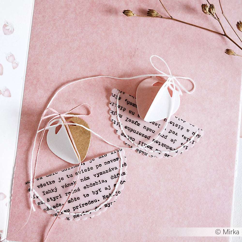 Zwei handgefertigte Engelsornamente aus Papier von Alexandra Renke mit Textflügeln und Schnurschleifen ruhen auf einem rosa Hintergrund.