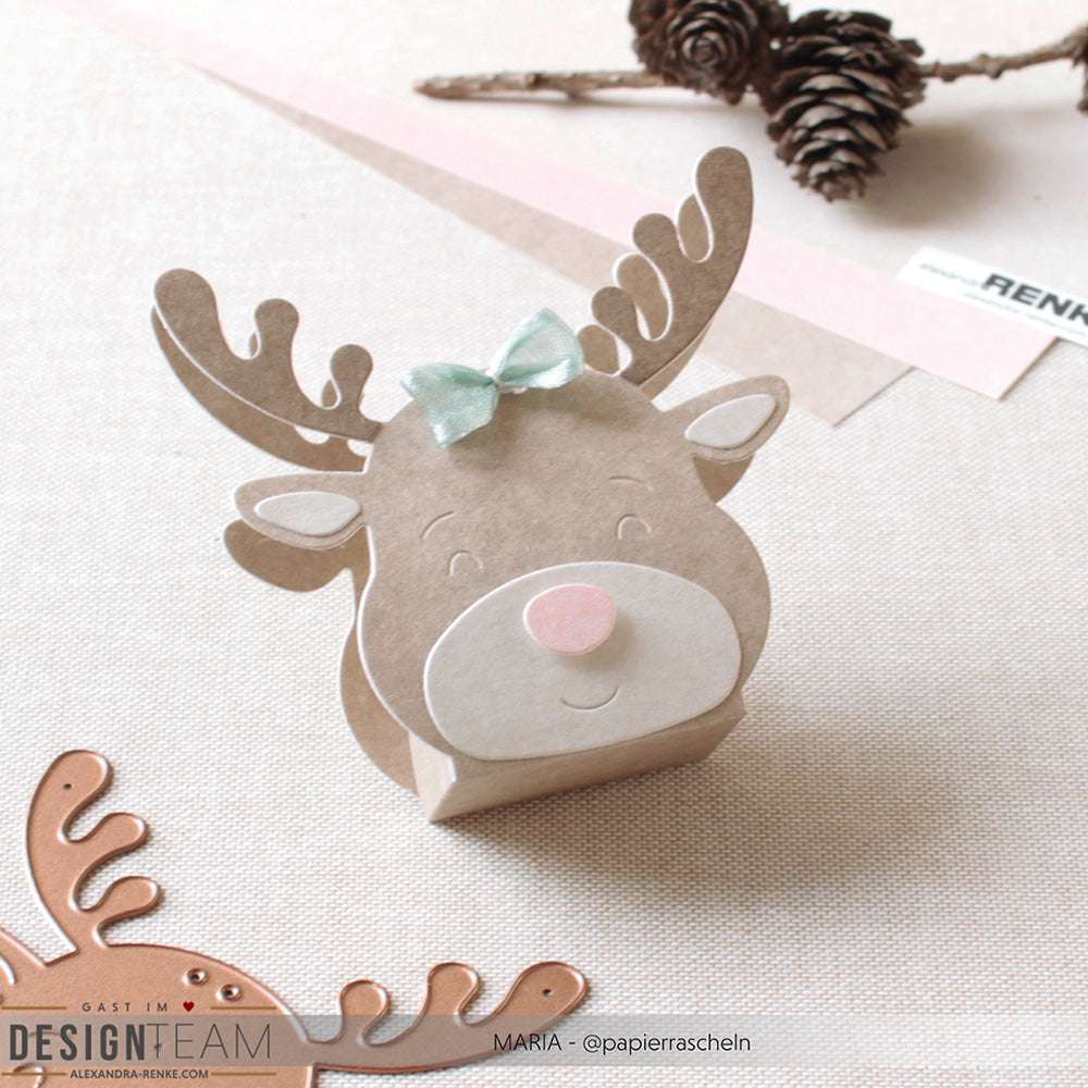 Ein Rentier in Form eines Papiers mit Alexandra Renke Designpapieren und einer mintgrünen Schleife, die mit der Motivstanze Rentier-Box hergestellt wurde. Auf einer hellen Fläche mit Tannenzapfen in der Nähe arrangiert.