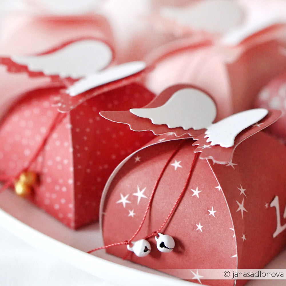 Rote Geschenkschachteln von Alexandra Renke aus Designpapier, verziert mit Engelsflügeln und Glöckchen, schön arrangiert auf einem weißen Teller.
