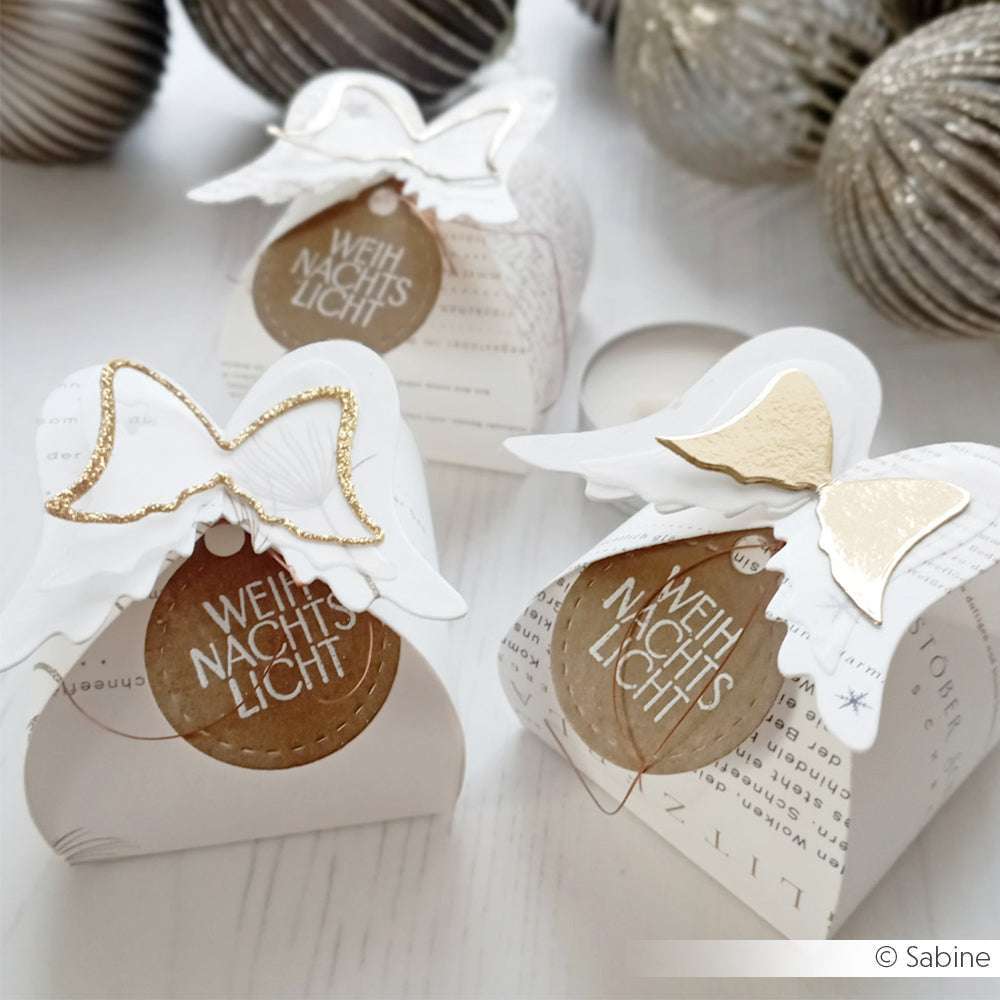 Drei weihnachtliche Geschenkboxen in Weiß und Gold von Alexandra Renke, hergestellt mit Designpapieren und dekoriert mit der Engelsflügel-Box-Stanze, zeigen Engelsflügel-Akzente und deutsche Textschilder auf weißer Oberfläche.