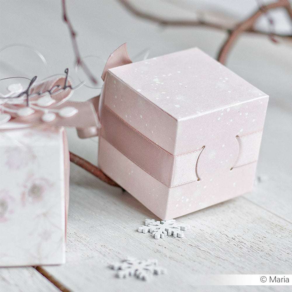 Eine zartrosa Geschenkbox mit Schleife, die mit Alexandra Renke Designpapieren und der Motivstanze Band-Box gestaltet wurde, ruht auf einer weißen Holzfläche neben einer Schneeflocken-Dekoration.