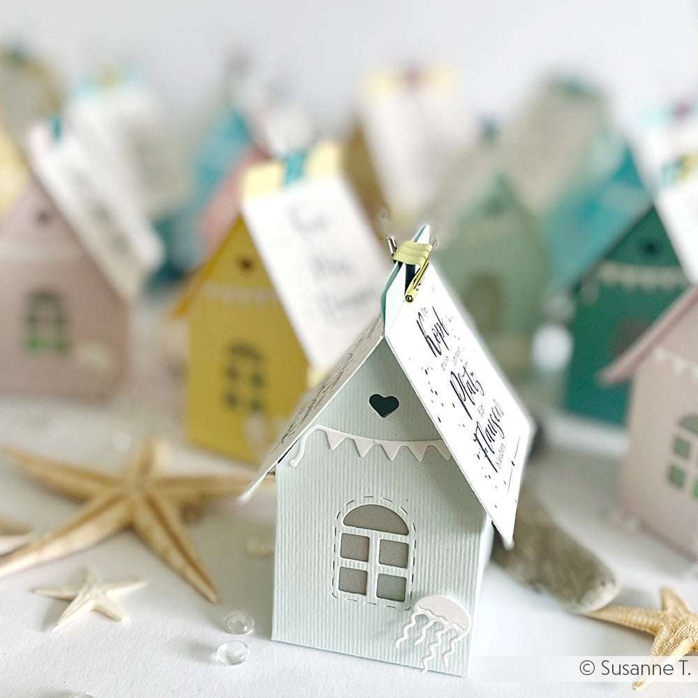 Pastellfarbene Papierhäuschen aus der Alexandra Renke Motivstanze Großes Häuschen, verziert mit dekorativen Anhängern, Muscheln und Seesternen, sind kunstvoll auf einer weißen Fläche angeordnet.