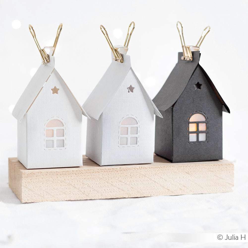 Drei kleine Papierhäuschen, zwei weiße und ein schwarzes, die mit Alexandra Renkes Motivstanze Kleines Häuschen aus edlem Designpapier gebastelt wurden, sitzen auf einem Holzblock, der mit goldenen Klammern verziert ist.