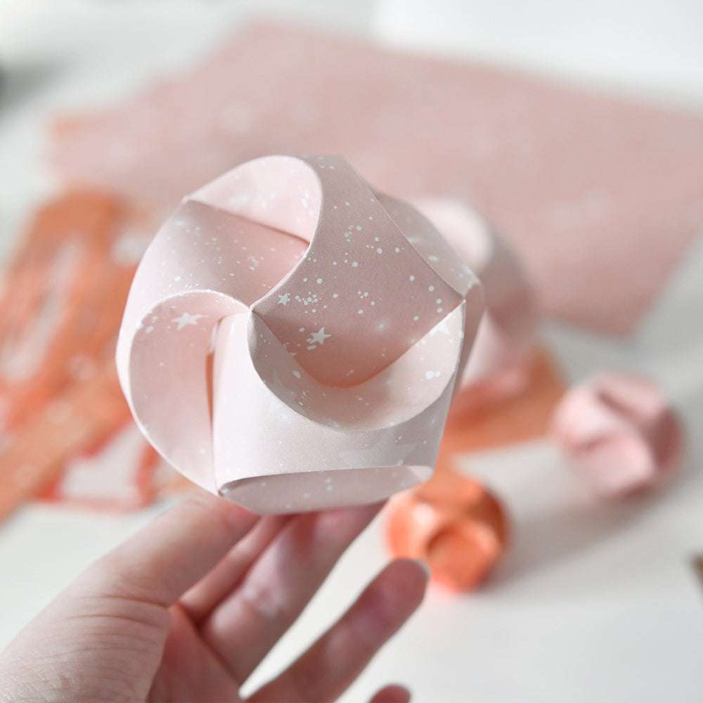 Eine Hand hält eine blassrosa, gesprenkelte Origami-Kugel, die mit der 3D-Dekokugel-Stanze von Alexandra Renke hergestellt wurde, während im Hintergrund weitere Papierkugeln verschwimmen.