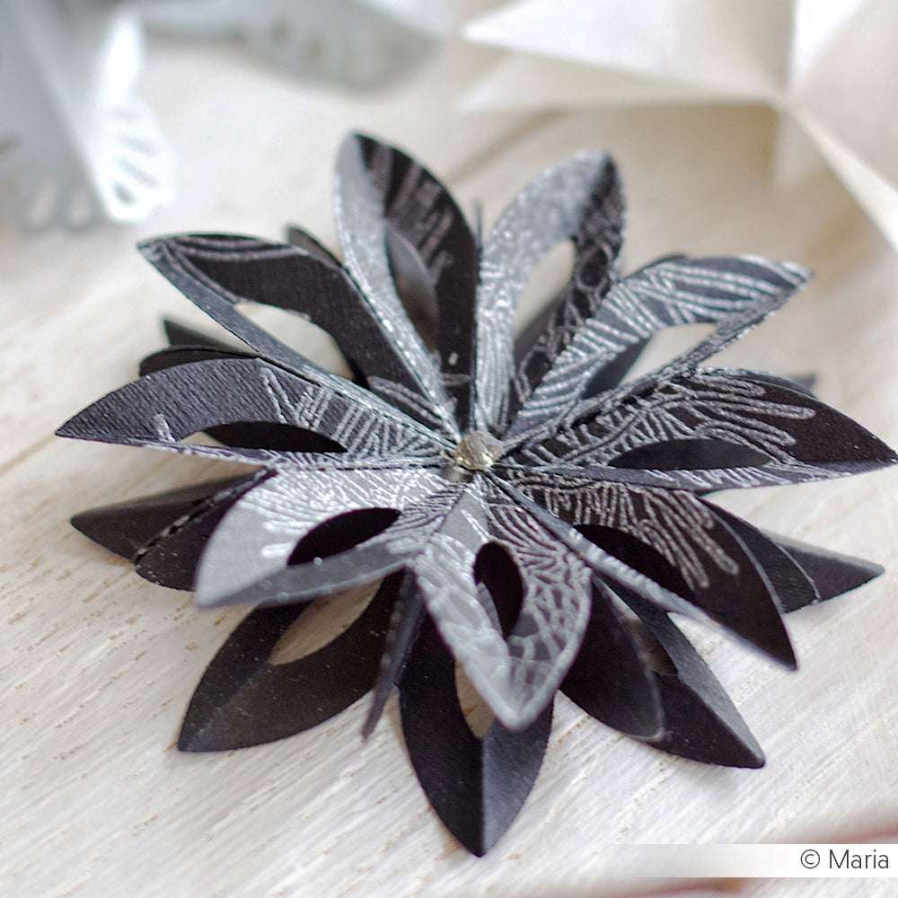 Eine schwarze und silberne Papierblume mit strukturierten Blütenblättern, die mit Alexandra Renke Designpapieren und DieCut Systemen hergestellt wurde, ruht auf einer hellen Holzoberfläche.