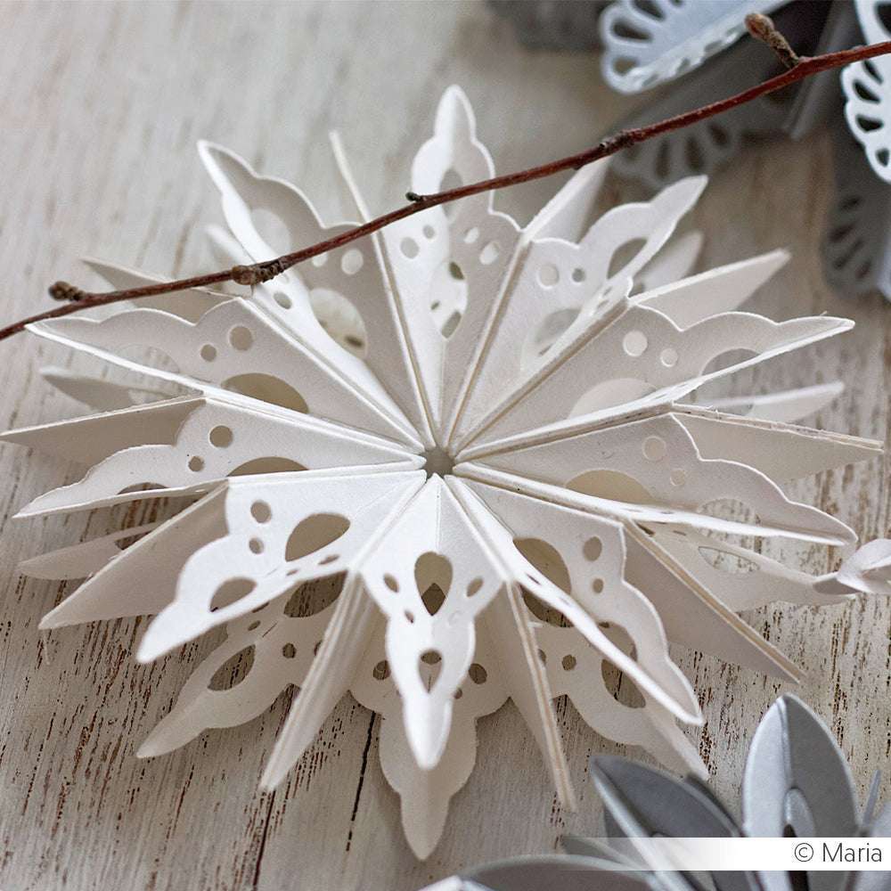 Eine weiße Schneeflocke aus Papier mit komplizierten Ausschnitten, die mit Alexandra Renke DieCut Systemen und Designpapieren hergestellt wurde, liegt auf einer Holzfläche neben einem Zweig.