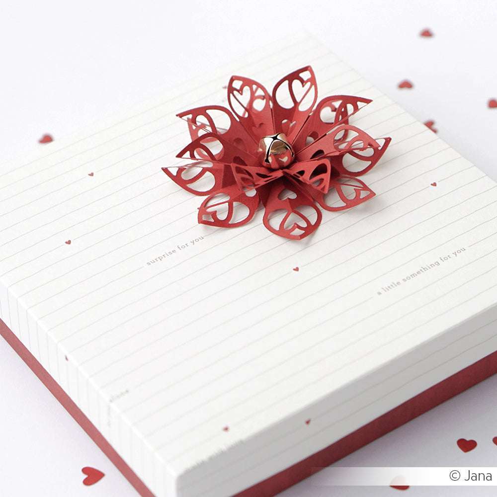 Ein weißer Geschenkkarton mit einer roten, herzförmigen Papierblume aus der Motivstanze Sternsegment Herzform von Alexandra Renke, gekrönt von einem Glöckchen und umgeben von kleinen roten Herzen aus hochwertigen Designpapieren.