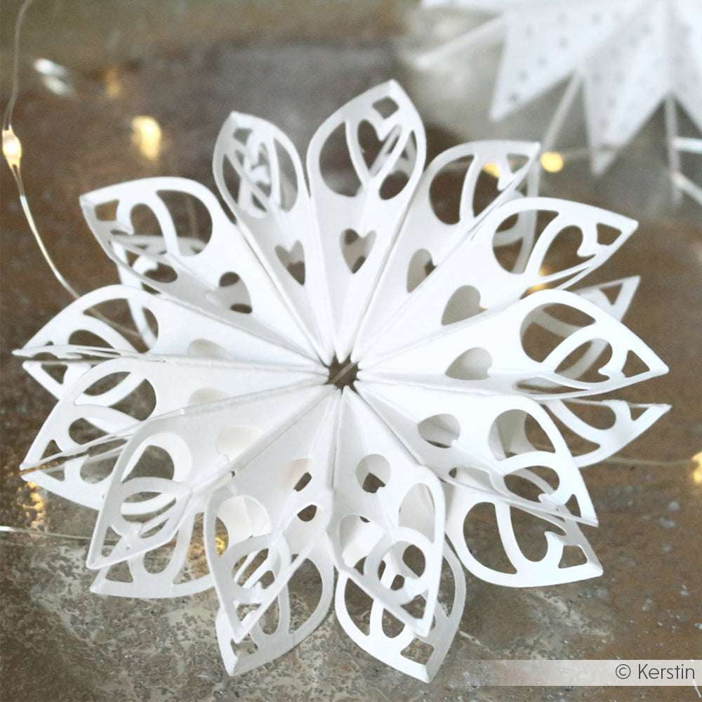 Ein kunstvoll geschnittenes Schneeflockenornament aus weißem Papier von Alexandra Renke, das mit einer Sternsegment Herzform Stanze hergestellt wurde, ruht auf einer reflektierenden Oberfläche mit sanften Lichtern im Hintergrund.