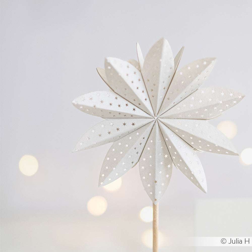 Eine weiße, sternförmige Papierdekoration von Alexandra Renke, hergestellt mit Designpapieren und einer Motivstanze Sternsegment, mit kleinen silbernen Punkten auf einem Stab vor einem verschwommenen hellen Hintergrund.