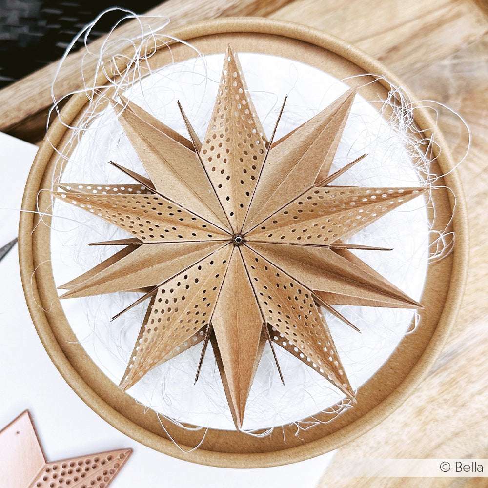 Ein hölzernes, gelochtes Sternornament von Alexandra Renke - gefertigt mit einem Motivstanze-Sternsegment - steht auf einem runden weißen Sockel, der mit weißen Fäden verziert ist.