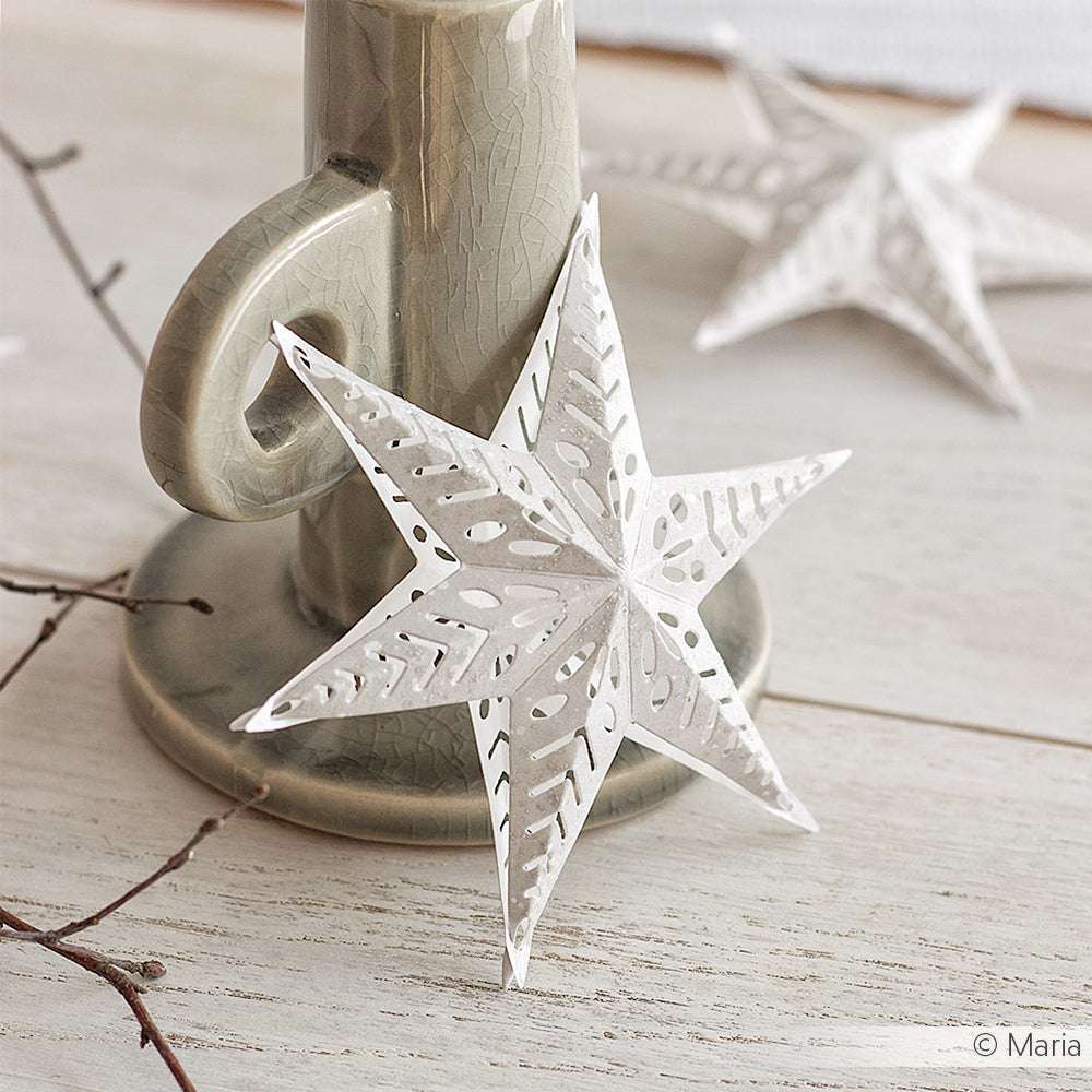 Der weiße Papierstern von Alexandra Renke, der mit DieCut Systemen und Motivstanze Steckstern hergestellt wurde, zeigt filigrane Ausschneidedetails, während er auf einem Kerzenhalter aus Keramik auf einer hellen Holzoberfläche ruht.