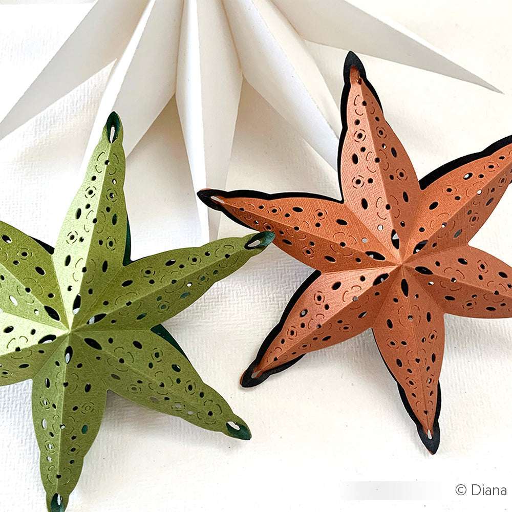 Zwei Papierstern-Ornamente, grün und braun, mit der Alexandra Renke Motivstanze Steckstern hergestellt und mit detaillierten Ausschneidemustern versehen, ruhen auf einem weißen Hintergrund.