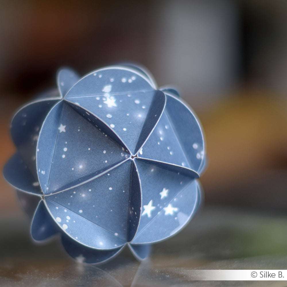 Eine blaue Origami-Kugel mit Sternmuster, die von Alexandra Renke Designpapiere mit einer Motivstanze hergestellt wurde, ruht auf einer unscharfen Fläche.