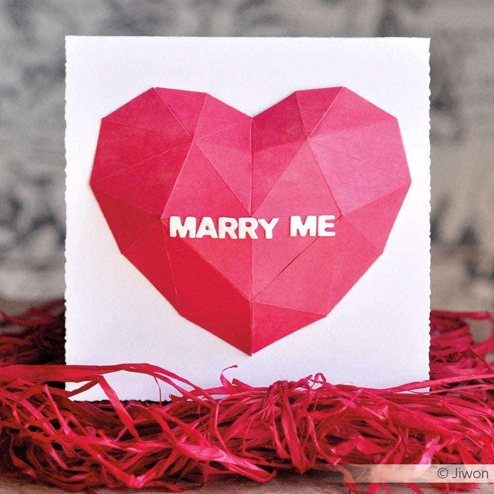 Ein rotes Origami-Herz mit "MARRY ME" in Motivstanze Blockbuchstaben sitzt auf einer weißen Karte auf rotem Schmuckpapier. Marke: Alexandra Renke.