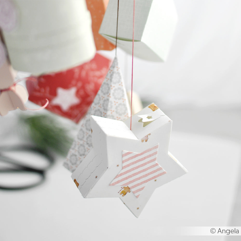 Alexandra Renke Papierornamente, wie eine sternförmige Box mit rosa Streifen aus Designpapieren und der Motivstanze Abriss-Stern Box DieCut Systeme, hängen anmutig an Schnüren vor einem hellen Hintergrund.