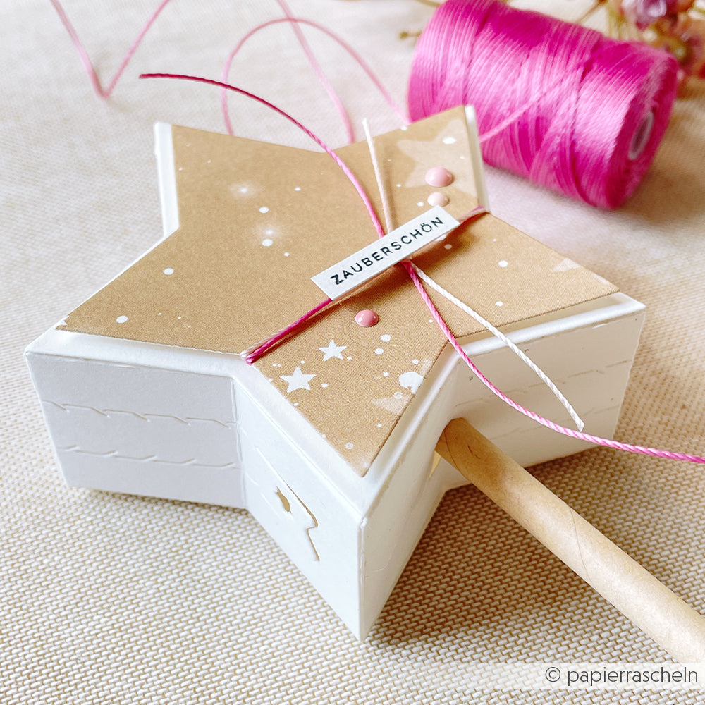 Die sternförmige, weiß-braune Geschenkbox mit Alexandra Renke Designpapier und rosafarbenem Garn mit der Aufschrift Zauberschön steht neben einer rosafarbenen Fadenspule - perfekt hergestellt mit dem Abriss-Stern Box DieCut System.