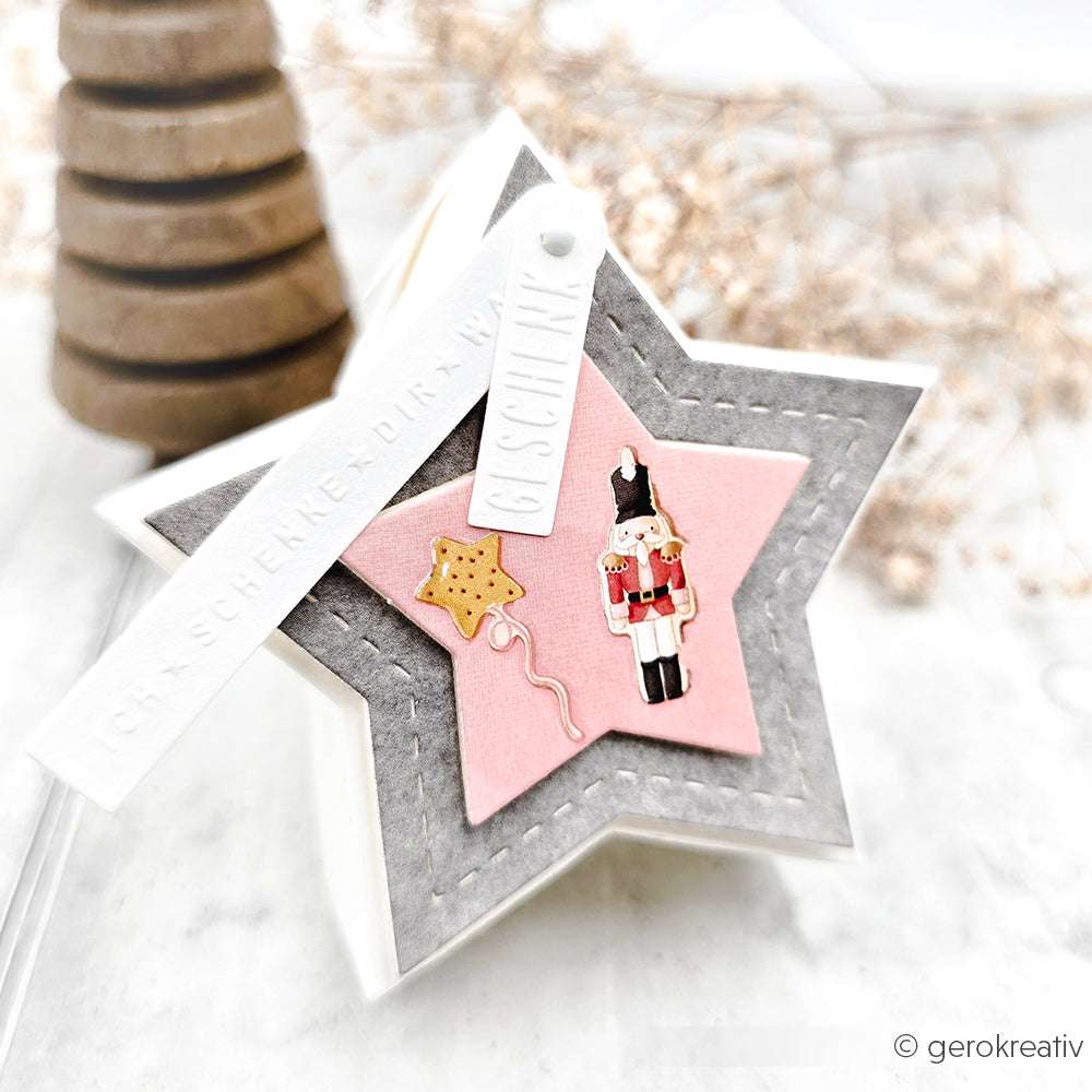 Sternförmige Geschenkschachtel mit rosa und grauen Lagen von Alexandra Renke, gefertigt mit der Motivstanze Abriss-Stern Box, dekoriert mit Nussknacker und Sternkeksen, Designpapieren und mit weißen Geschenkanhängern versehen.