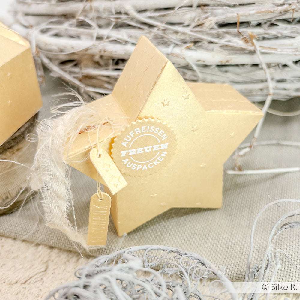 Goldfarbene Geschenkbox in Form eines Sterns, hergestellt mit der Motivstanze Abriss-Stern Box von Alexandra Renke, versehen mit einem weißen Siegel, einem Stoffband und einem Anhänger auf einer neutral strukturierten Oberfläche - ideal, um Ihre Stanzformen oder Stanzsysteme zu präsentieren.