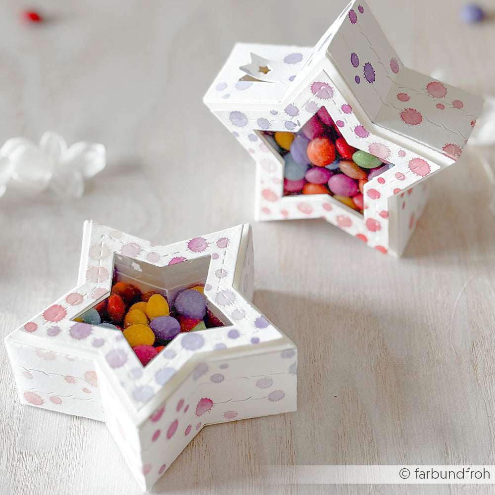 Zwei sternförmige Schachteln von Alexandra Renke, die mit einer Stanzsysteme Motivstanze hergestellt und mit Tupfen verziert wurden, sind mit bunten Pralinen gefüllt und werden auf einer hellen Fläche ausgestellt.