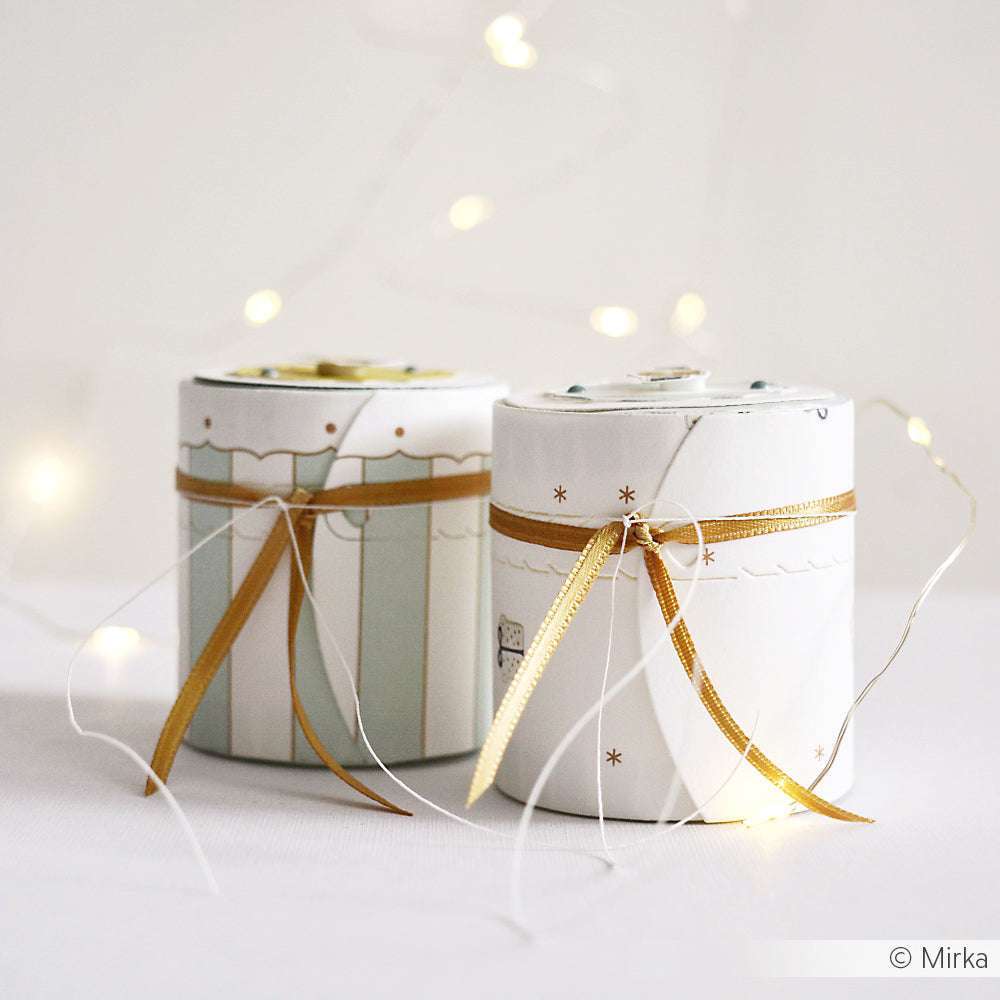 Zwei Toilettenpapierrollen, die mit Bändern und goldenen Akzenten verziert sind und mit Techniken von Alexandra Renke hergestellt wurden, werden mit Lichterketten im Hintergrund ausgestellt, die für einen festlichen Touch sorgen.