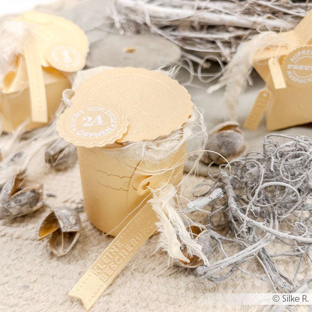 Kleine beigefarbene Geschenkboxen mit strukturierten Anhängern, hergestellt mit Alexandra Renke Motivstanze Abriss-Zylinder Box und Stanzsystemen, verziert mit natürlichen Elementen auf einer leicht rustikalen Oberfläche.