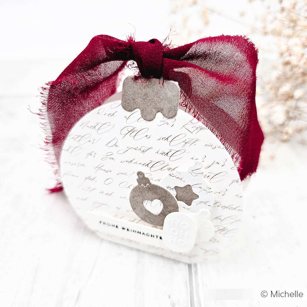 Eine runde, handgeschriebene Geschenkbox von Alexandra Renke mit burgunderfarbener Stoffschleife und weihnachtlichen Ausschneideverzierungen, hergestellt mit Dies und Stanzsystemen wie der Motivstanze Baumkugel Box.
