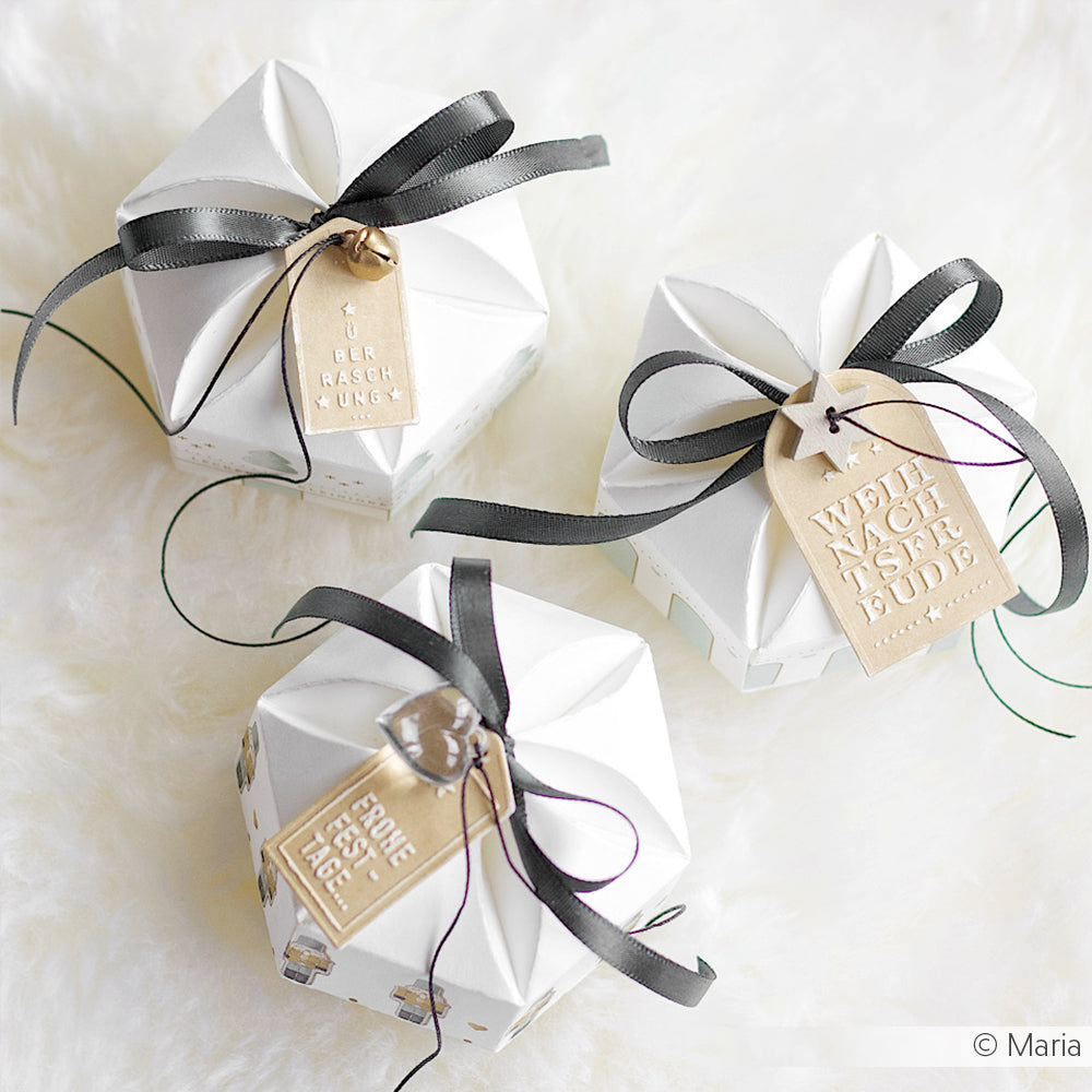 Drei weiße, geometrische Geschenkboxen mit schwarzen Bändern und braunen Anhängern, hergestellt von Alexandra Renke Designpapieren, werden auf einer weichen weißen Oberfläche präsentiert. Ideal für kreative Projekte mit Motivstanze Box mit Blütenverschluss oder Stanzsystemen.