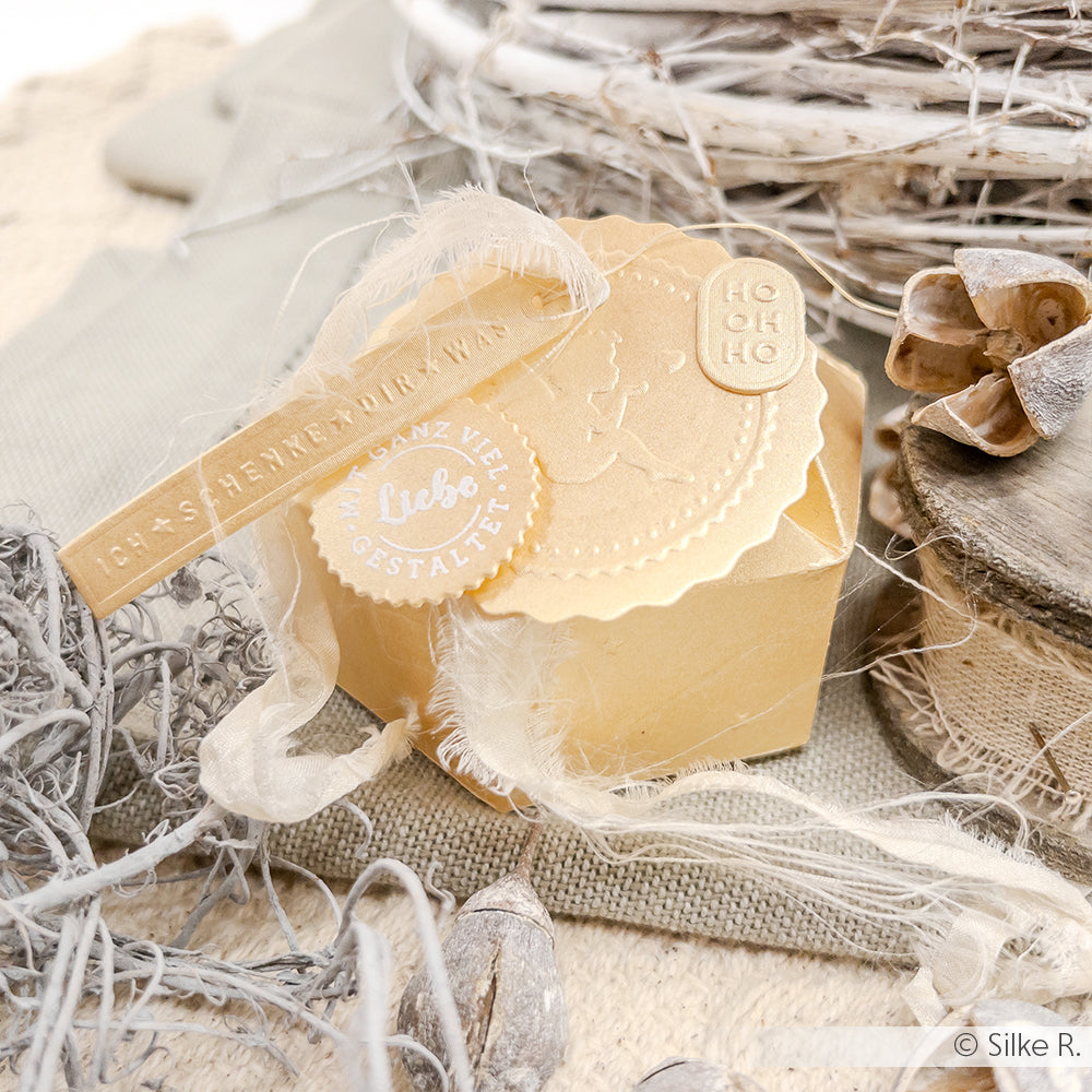 Goldfarbene Geschenkbox von Alexandra Renke mit dekorativem Band, Feder und geprägten Anhängern, gefertigt aus eleganten Designpapieren, platziert auf neutralem, rustikalem Stoff und Zweigen.