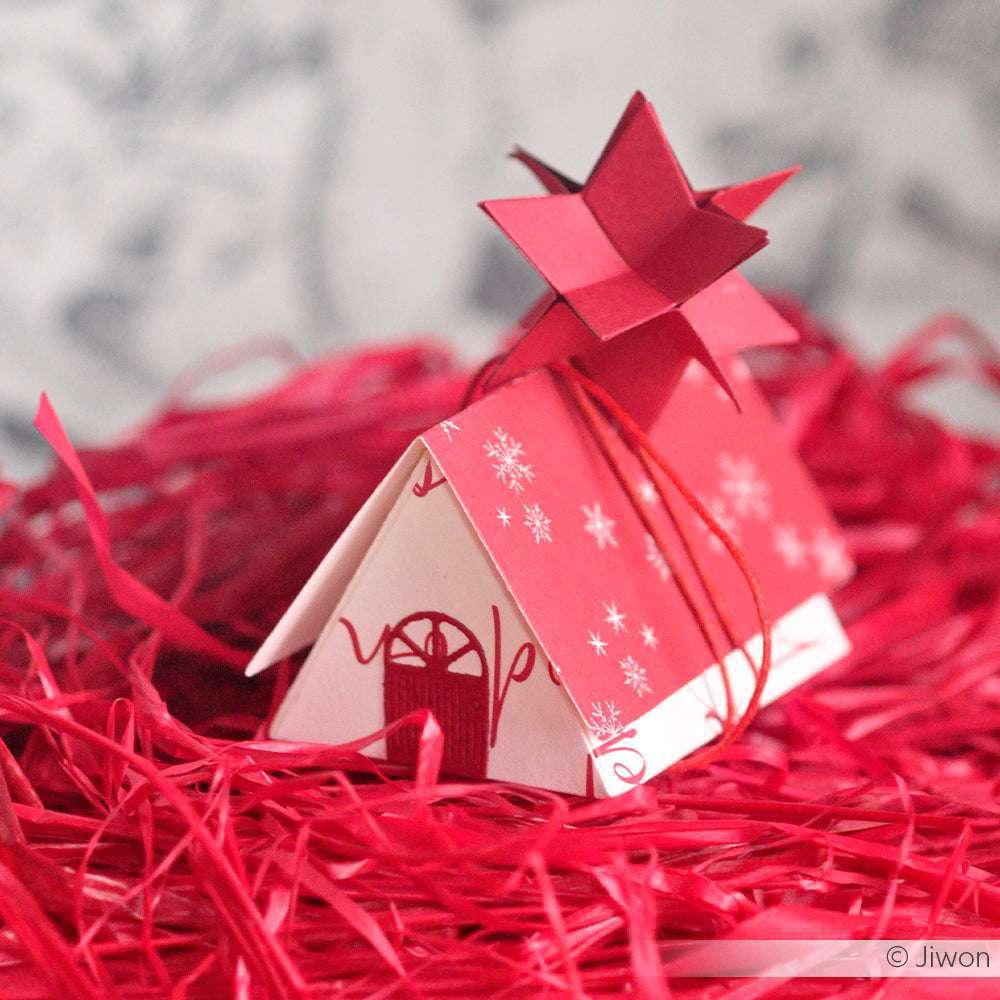 Ein kleines rot-weißes Papierhaus mit einem Stern auf der Spitze, gefertigt aus Alexandra Renke Designpapieren, sitzt auf geschreddertem rotem Papier - eine charmante Inspiration für Projekte von Motivstanze Dreieck Klappbox oder DieCut Systeme.