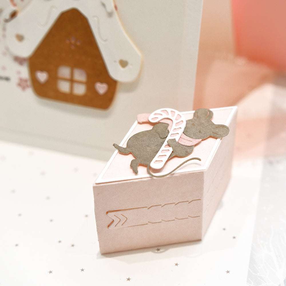 Eine rosafarbene Geschenkbox von Alexandra Renke, die mit Motivstanze gefertigt und mit einer Maus und einer Zuckerstange verziert ist, steht neben einer Karte mit einem Lebkuchenhausmotiv.