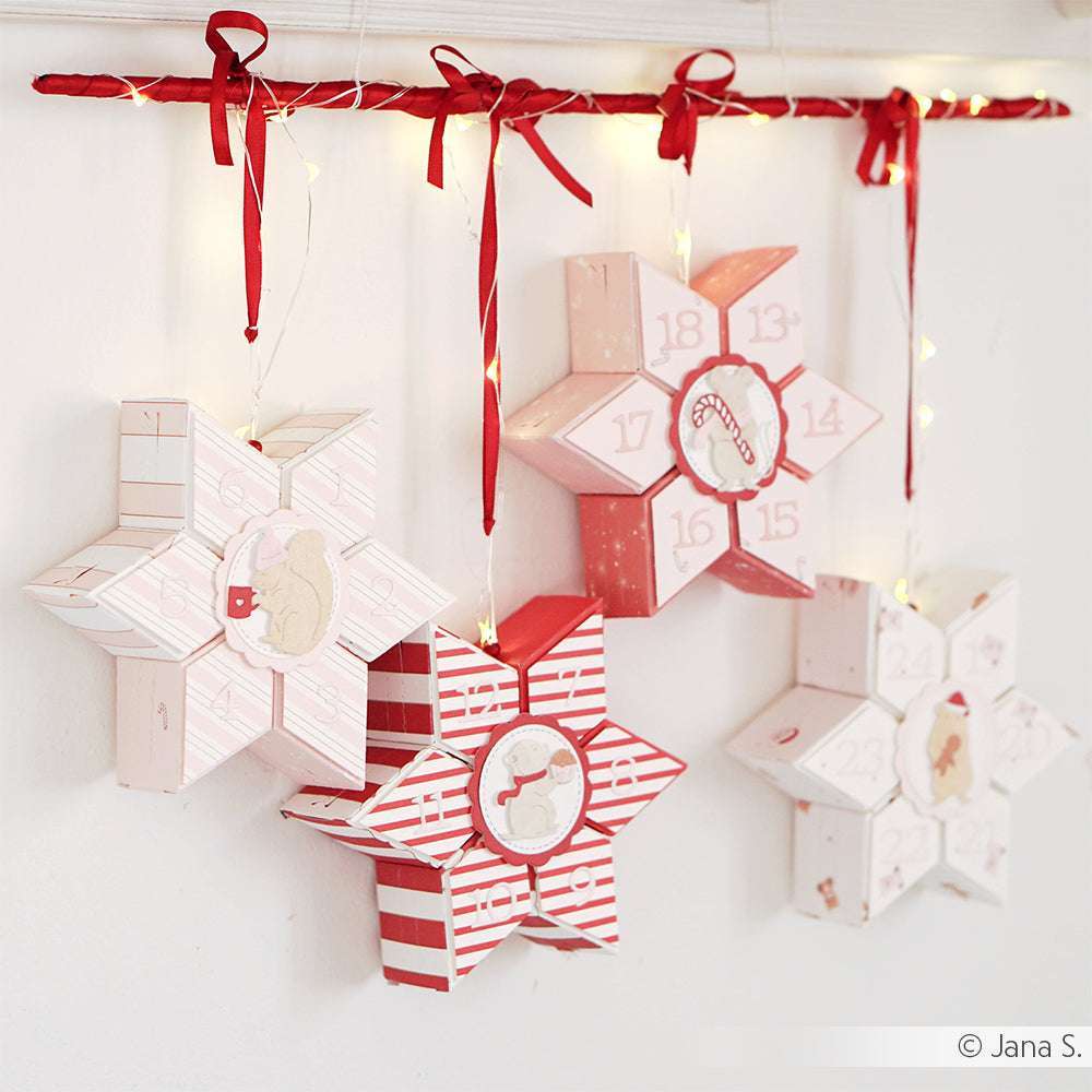 Vier rot-weiße Adventskalender in Sternform von Alexandra Renke, gestaltet mit DieCut Systemen, hängen an einer Stange mit roten Bändern und funkelnden Lichterketten.