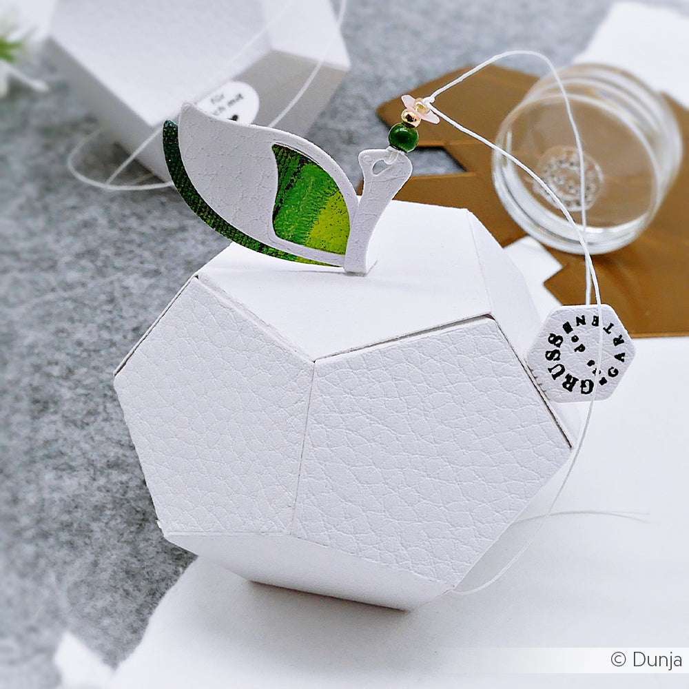 Geometrische weiße Geschenkbox in Form eines Apfels mit grünem Blatt, hergestellt mit der Motivstanze Apfel Box von Alexandra Renke und Stanzsystemen, mit einem kleinen Anhänger, der an einer Schnur befestigt ist.
