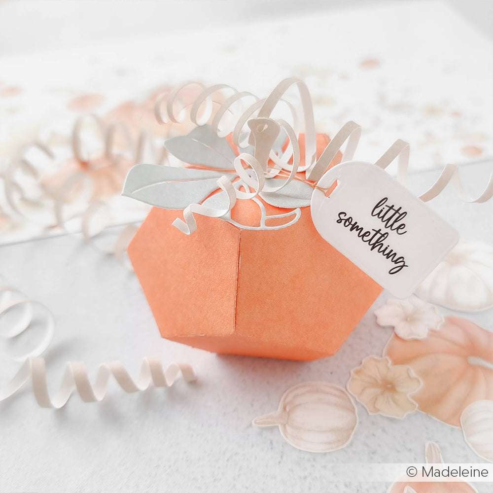 Orangefarbene Papierkürbis-Geschenkbox, hergestellt mit der Apfel Box und den DieCut Systemen von Alexandra Renke, verziert mit einem gewellten Band und einem "Kleinigkeit"-Anhänger, dargestellt auf einem pastellfarbenen Hintergrund.