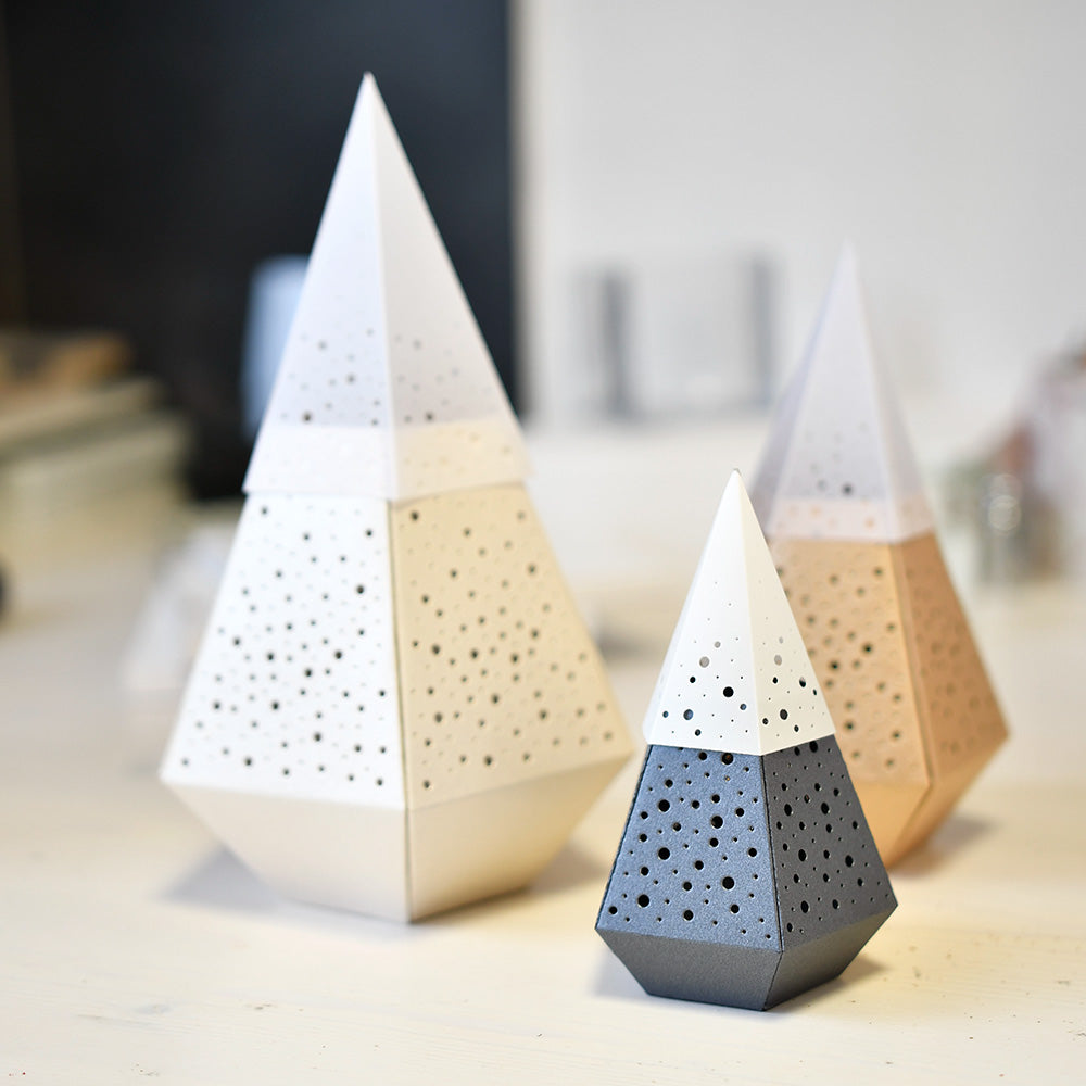 Drei geometrische, gelochte Kegelskulpturen von Alexandra Renke - weiß, beige und grau -, die mit Deep Edge Motivstanzen gefertigt wurden, werden kunstvoll auf einem Leuchttisch präsentiert.