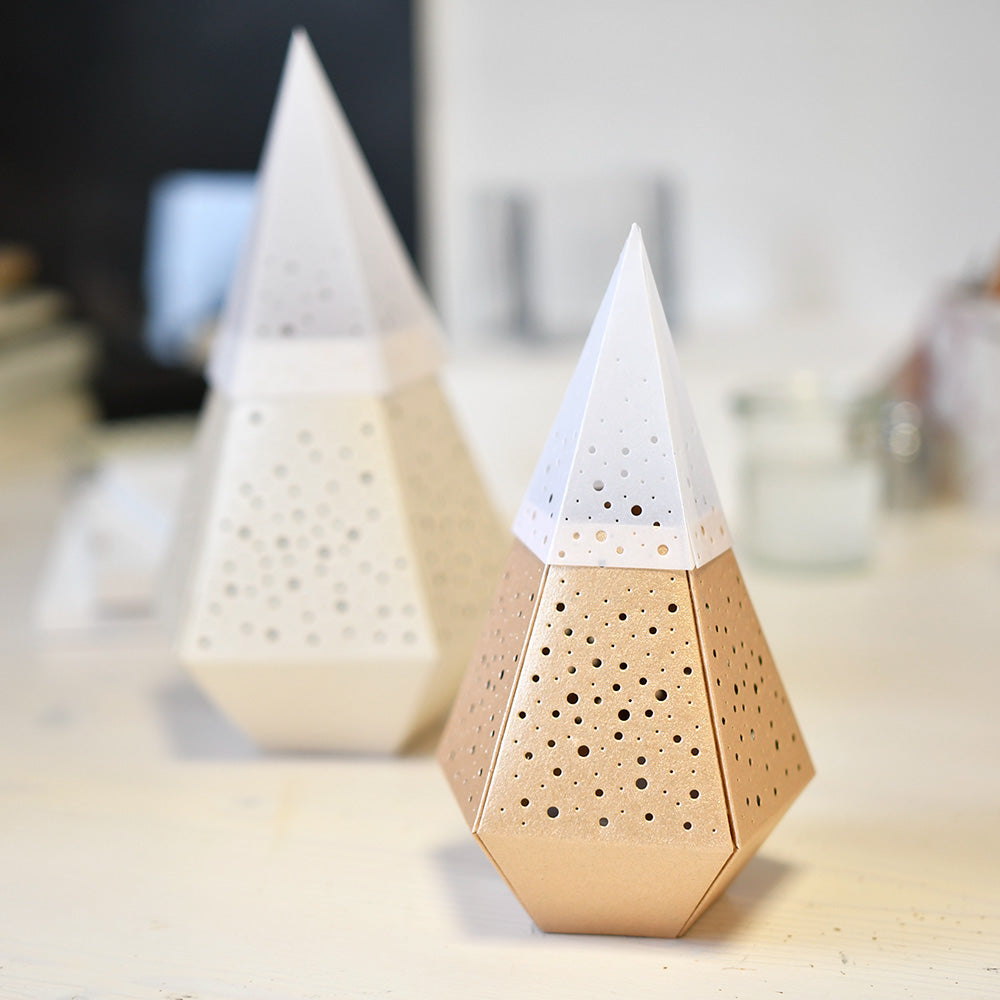 Zwei geometrische, kegelförmige Objekte mit perforierten Löchern - hergestellt mit Alexandra Renkes PAPPcut®ies Motivstanze Windlicht - ein goldenes und ein weißes, ausgestellt auf einer hellen Fläche.