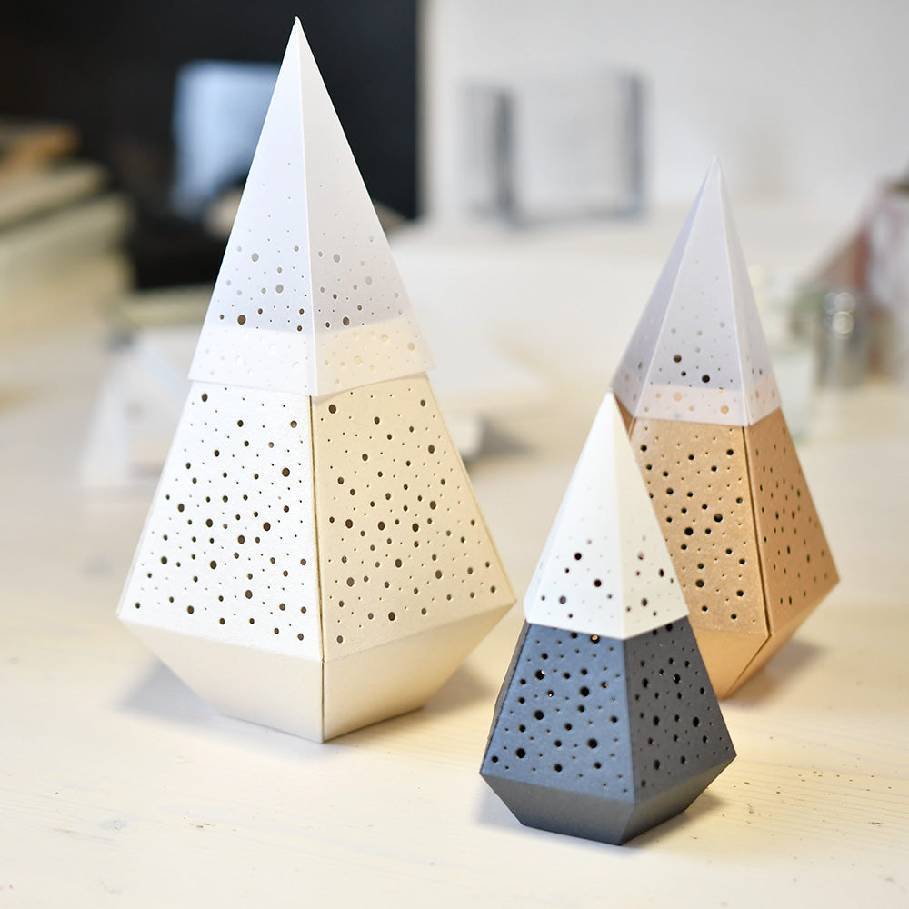 Drei geometrische, kegelförmige Papierlaternen von Alexandra Renke, die mit PAPPcut®ies und Motivstanze Windlicht gefertigt wurden und kleine Löcher in neutralen Farben aufweisen, ruhen auf einer hellen Fläche.