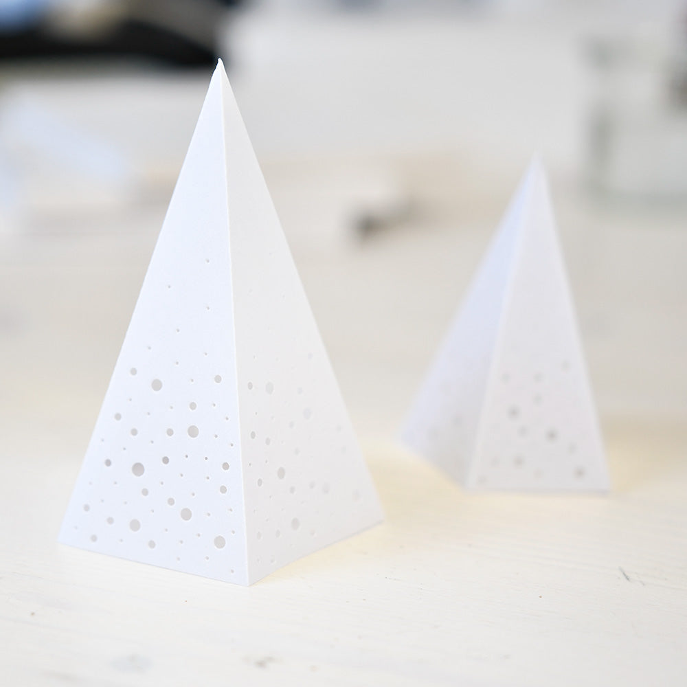 Zwei weiße Papierpyramiden mit kleinen kreisförmigen Löchern, die mit Alexandra Renke Deep Edge Motivstanzen hergestellt wurden, werden auf eine helle Fläche gelegt.