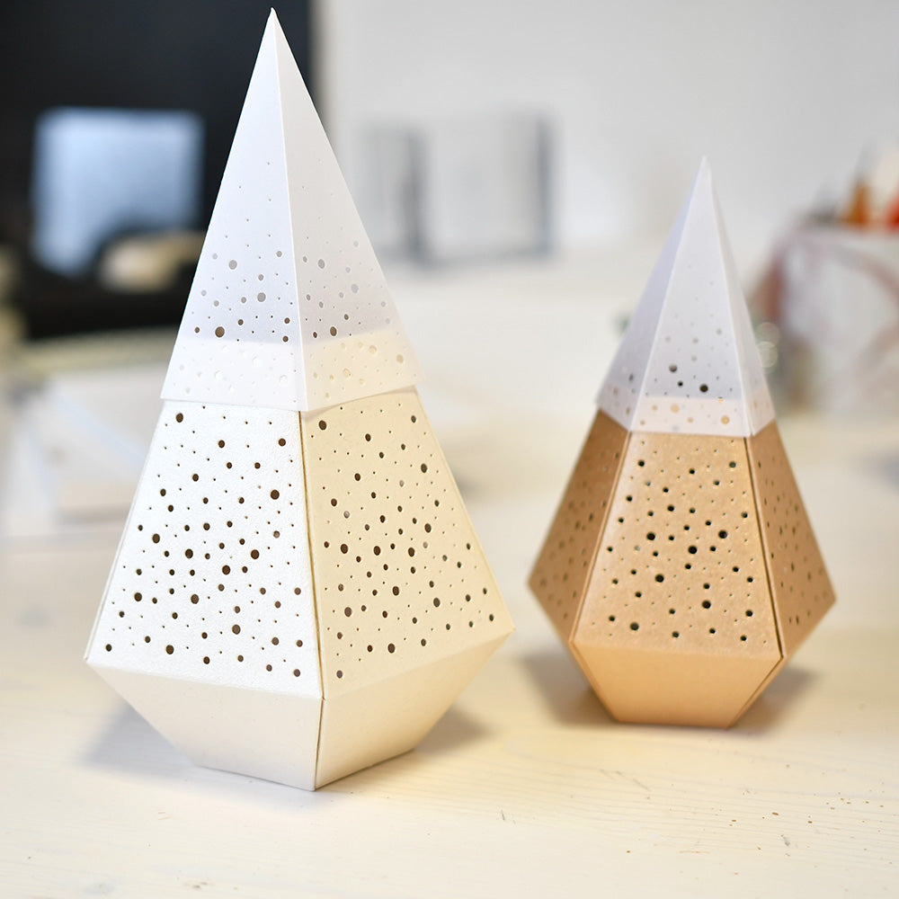 Zwei geometrische, pyramidenförmige Windlichter von Alexandra Renke verleihen einem hellen Tisch einen besonderen Charme. Ihre perforierten Muster werfen komplizierte Schatten.