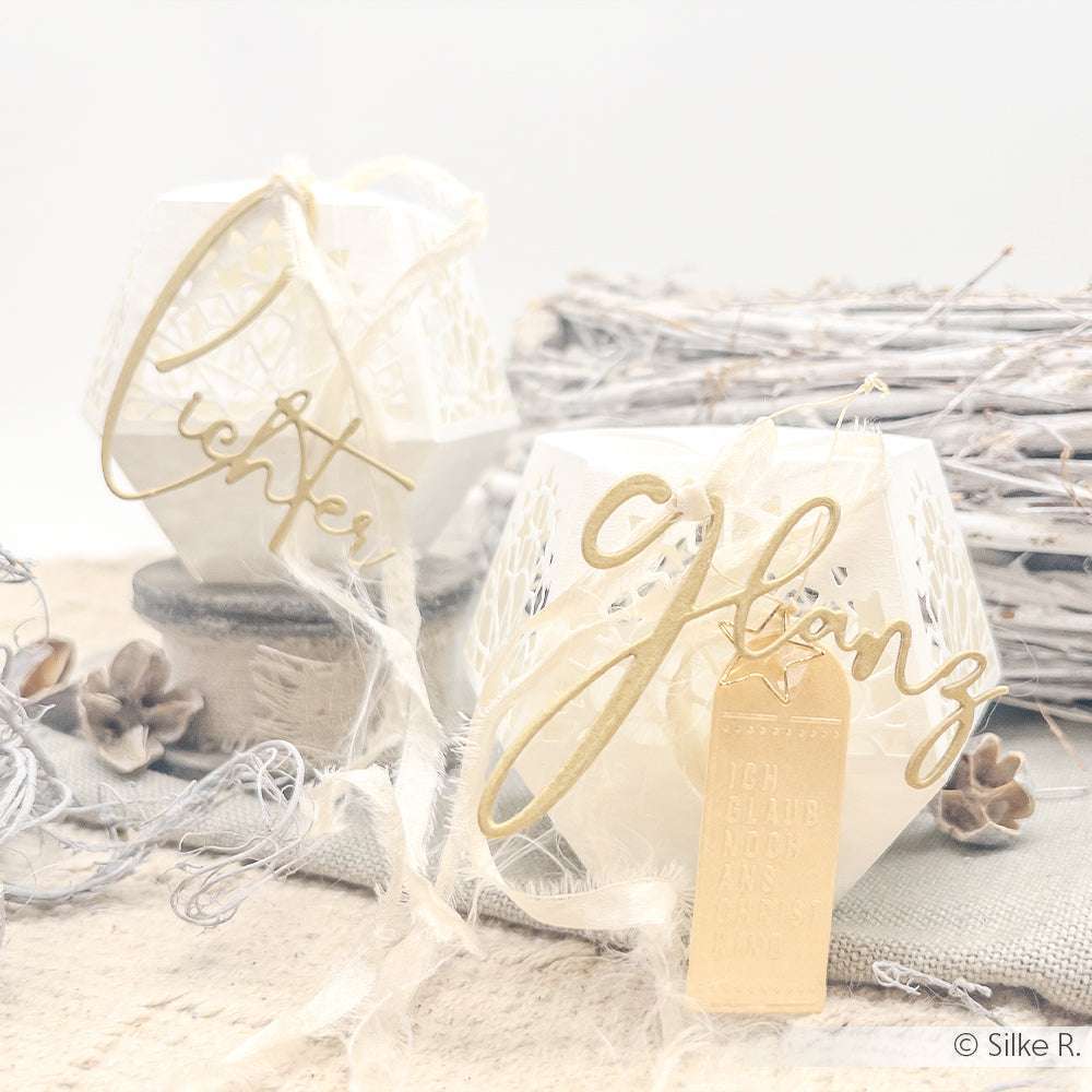 Zwei weiße Laternen von Alexandra Renke mit goldenen Schriftanhängern, Spitzenbändern, Tannenzapfen, rustikalen Holzakzenten und PAPPcut®ies für mehr Charme.