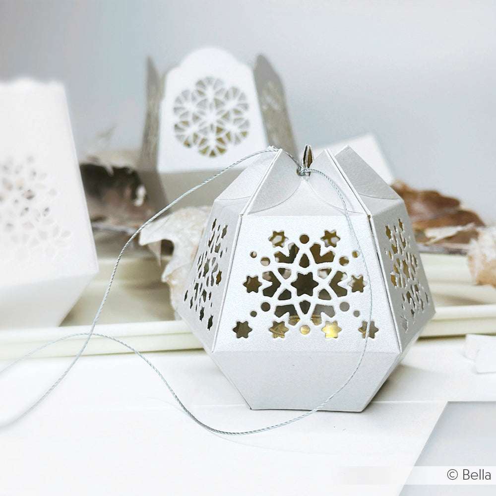 Eine silberne, geometrische Papierlaterne mit verschlungenen Sternausschnitten, hergestellt mit Deep Edge Motivstanzen von Alexandra Renke, steht auf einer weißen Fläche und ist mit einer Schnur befestigt.