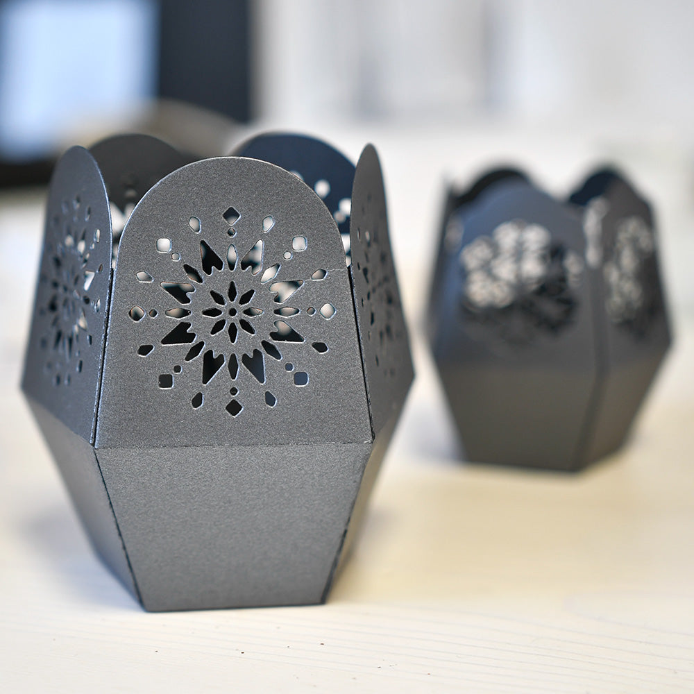 Dunkelgraue Metall-Kerzenhalter von Alexandra Renke mit aufwendigen Windlicht-Ornamenten, die auf einer hellen Oberfläche angebracht sind.